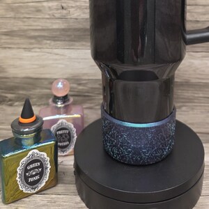 Puede incluir: Un termo de viaje Stanley negro y dorado con asa y pajita. El termo tiene el logotipo de Stanley y una banda dorada. El termo est&aacute; sobre un soporte negro, con dos frascos de perfume decorativos en el fondo.