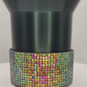 Puede incluir: Un vaso de color gris oscuro con una banda de pedrería cuadrada iridiscente. La pedrería refleja un arcoíris de colores, incluyendo rosa, verde y amarillo. El vaso tiene un diseño elegante y moderno.