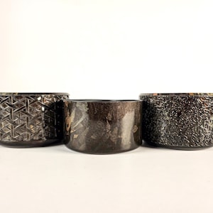 May include: Three cylindrical containers with dark, metallic finishes. The left container features a woven pattern, the center one has a smooth, mottled dark surface, and the right container displays an intricate, embossed floral-like texture.