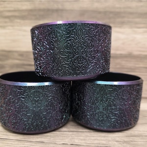 Puede incluir: Tres cuencos decorativos iridiscentes apilados. Los cuencos tienen un exterior oscuro y texturizado con un patr&oacute;n ornamental en relieve. Los bordes tienen un sutil brillo p&uacute;rpura y azul. Cada cuenco mide unos 8 cm de di&aacute;metro.