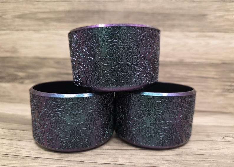 Puede incluir: Tres cuencos decorativos iridiscentes apilados. Los cuencos tienen un exterior oscuro y texturizado con un patr&oacute;n ornamental en relieve. Los bordes tienen un sutil brillo p&uacute;rpura y azul. Cada cuenco mide unos 8 cm de di&aacute;metro.