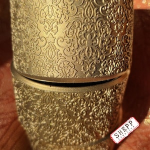 May include: A gold-colored, cylindrical container with an ornate, embossed pattern. The container is reflected in a mirrored surface. The container has a decorative, vintage-inspired design. The container is likely used for storage or display.