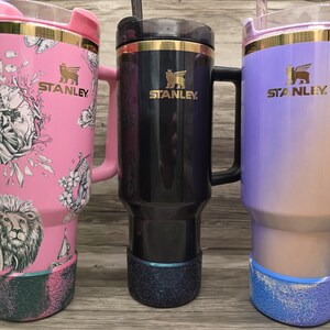 Puede incluir: Tres tazas de viaje Stanley con asas. Una es rosa con un dise&ntilde;o de le&oacute;n y flores, otra es negra y la tercera es un degradado de morado y beige. Todas tienen detalles dorados y el logotipo de Stanley.