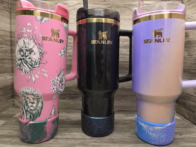Puede incluir: Tres tazas de viaje Stanley con asas. Una es rosa con un dise&ntilde;o de le&oacute;n y flores, otra es negra y la tercera es un degradado de morado y beige. Todas tienen detalles dorados y el logotipo de Stanley.
