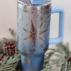 May include: A stainless steel tumbler with a blue lid and handle. The tumbler has a glitter finish and a snowflake design.