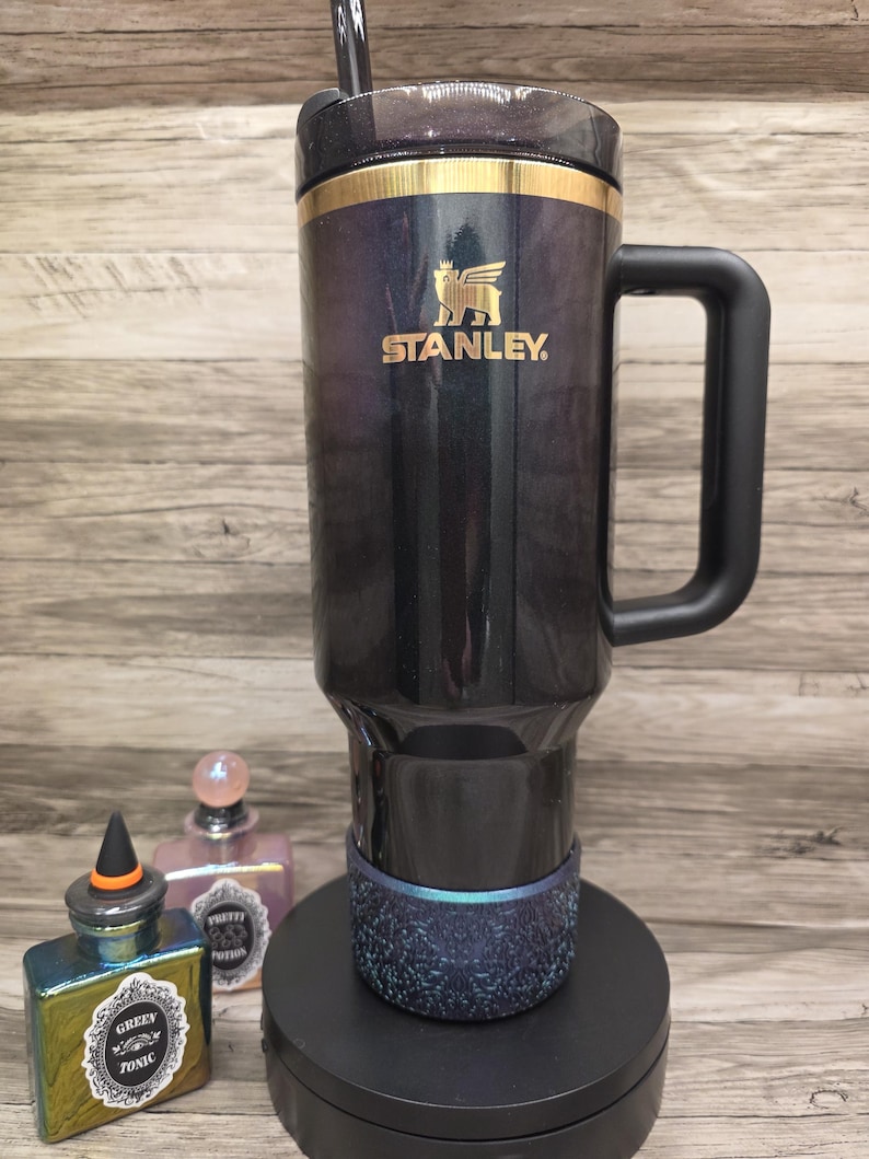 Puede incluir: Un termo de viaje Stanley negro y dorado con asa y pajita. El termo tiene el logotipo de Stanley y una banda dorada. El termo est&aacute; sobre un soporte negro, con dos frascos de perfume decorativos en el fondo.