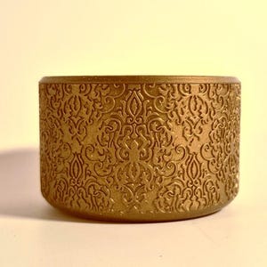 May include: A cylindrical, gold-colored container with an embossed, ornate pattern. The design covers the entire exterior surface of the container. The container is sitting on a light-colored surface.
