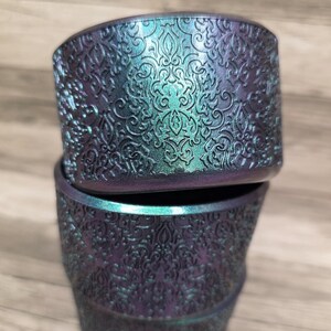 Puede incluir: Dos macetas apiladas, iridiscentes, con un detallado patr&oacute;n floral en relieve. Las macetas tienen un color base p&uacute;rpura oscuro con reflejos turquesa y verde. El dise&ntilde;o es un trabajo de volutas ornamentado y repetitivo.