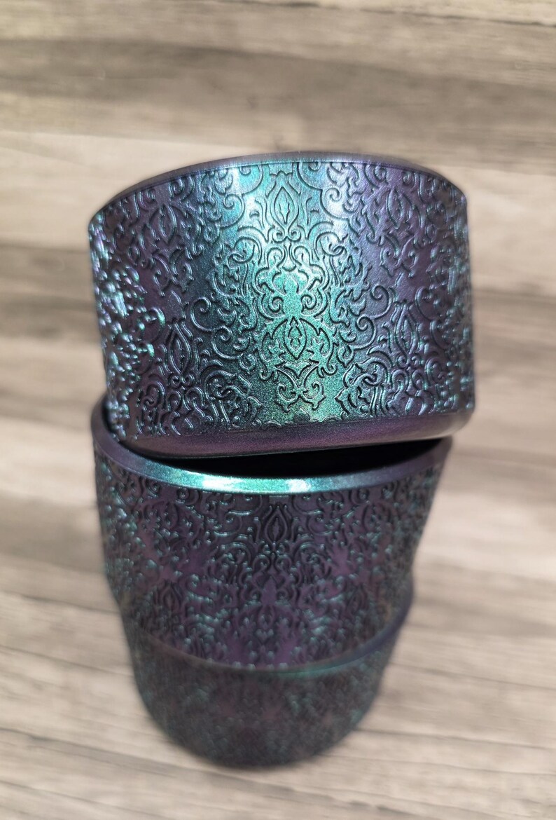 Puede incluir: Dos macetas apiladas, iridiscentes, con un detallado patr&oacute;n floral en relieve. Las macetas tienen un color base p&uacute;rpura oscuro con reflejos turquesa y verde. El dise&ntilde;o es un trabajo de volutas ornamentado y repetitivo.