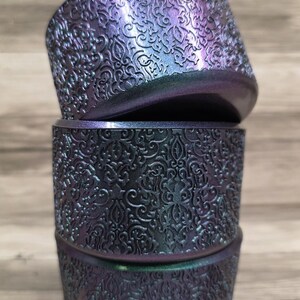 Puede incluir: Tres recipientes cil&iacute;ndricos apilados con un detallado patr&oacute;n floral en relieve. Los recipientes tienen un acabado que cambia de color, pasando del morado al verde azulado. Los recipientes son probablemente decorativos o para almacenamiento.