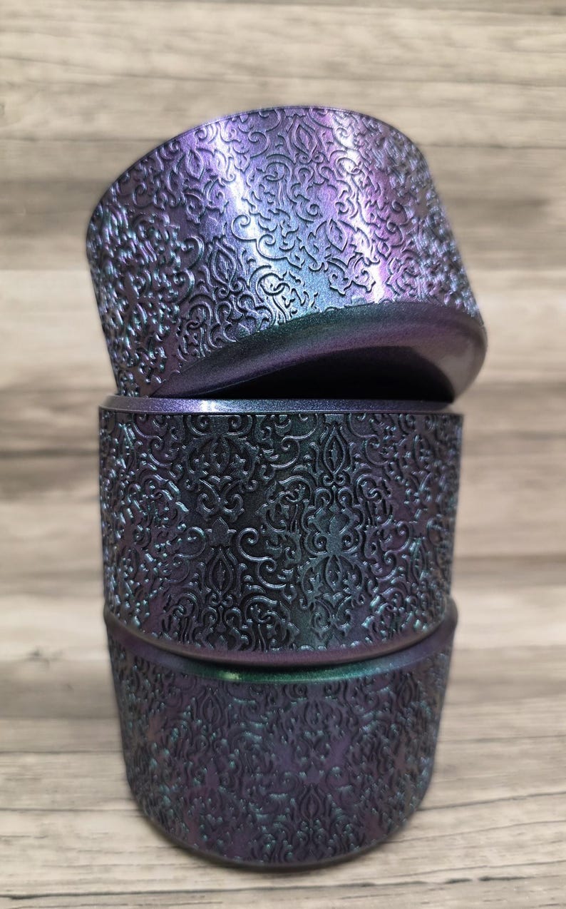 Puede incluir: Tres recipientes cil&iacute;ndricos apilados con un detallado patr&oacute;n floral en relieve. Los recipientes tienen un acabado que cambia de color, pasando del morado al verde azulado. Los recipientes son probablemente decorativos o para almacenamiento.