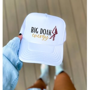May include: White trucker hat with the text "BIG DOAK energy" in black and gold lettering, and a maroon and white graphic. The hat has a white mesh back and a curved brim.