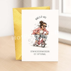 May include: A greeting card with a cartoon illustration of a woman riding a pink tricycle. The text "Hallo 40" and "Erwachsenwerden ist optional" are above and below the illustration. A gold envelope is behind the card.