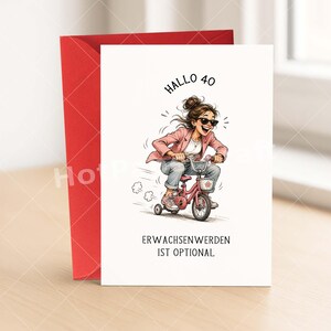 May include: A greeting card with a red envelope. The card features an illustration of a woman riding a pink tricycle. The text on the card reads "Hallo 40" and "Erwachsenwerden ist optional."