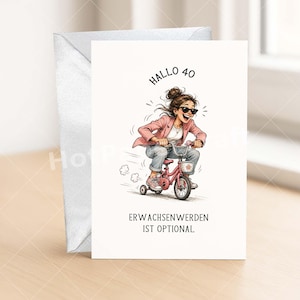 May include: A white greeting card with a cartoon illustration of a woman riding a pink tricycle. The card has the text "HALLO 40" and "ERWACHSENWERDEN IST OPTIONAL". A silver envelope is on the left.