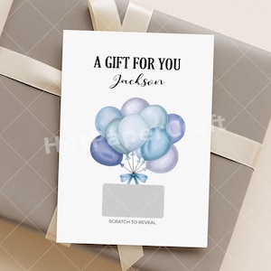 Könnte beinhalten: Eine weiße Geschenkkarte mit dem Text "A GIFT FOR YOU Jackson" und einer Gruppe blauer Luftballons. Unter den Luftballons befindet sich ein Rubbelbereich mit den Worten "SCRATCH TO REVEAL". Die Karte liegt auf einer grauen Geschenkbox.