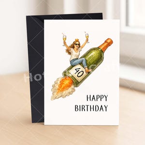 May include: A birthday card with a woman riding a champagne bottle rocket, holding two glasses of champagne. The card reads "Happy Birthday" with the number "40" on the bottle. A black envelope is on the left.