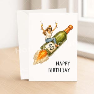 May include: A birthday card with a white envelope. The card features an illustration of a woman riding a champagne bottle rocket, holding two glasses of champagne. The bottle has the number 40 on it, and the words "HAPPY BIRTHDAY" are printed below.