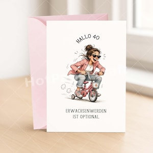 May include: A greeting card with a pink envelope. The card features an illustration of a woman riding a pink tricycle. The text on the card reads "Hallo 40" and "Erwachsenwerden ist optional."
