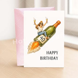 May include: A birthday card with a woman riding a champagne bottle rocket, holding two glasses of champagne. The bottle has a "40" label. The card reads "HAPPY BIRTHDAY" in black text. A pink envelope is behind the card.