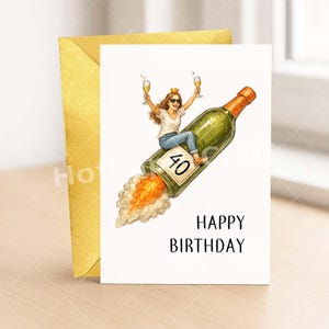 May include: A birthday card with a woman riding a champagne bottle rocket, holding two glasses of champagne. The card reads "Happy Birthday" and features the number "40". A gold envelope is behind the card.