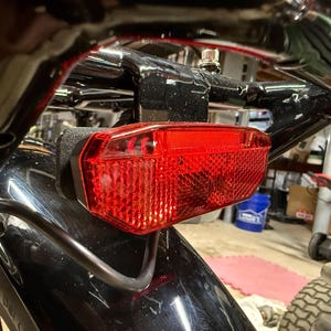 May include: Close-up of a red motorcycle tail light. The light is rectangular with a textured surface and is mounted on a black metal frame. The image is taken from a low angle, showing the light's details.