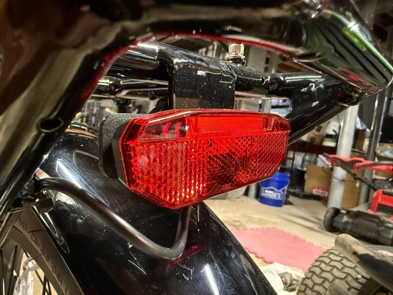 May include: Close-up of a red motorcycle tail light. The light is rectangular with a textured surface and is mounted on a black metal frame. The image is taken from a low angle, showing the light's details.
