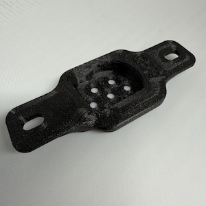 May include: A black 3D-printed bracket with a rectangular body and rounded ends. It features two holes at the ends and a recessed area with five smaller holes. The text "ASD" is visible on the side.