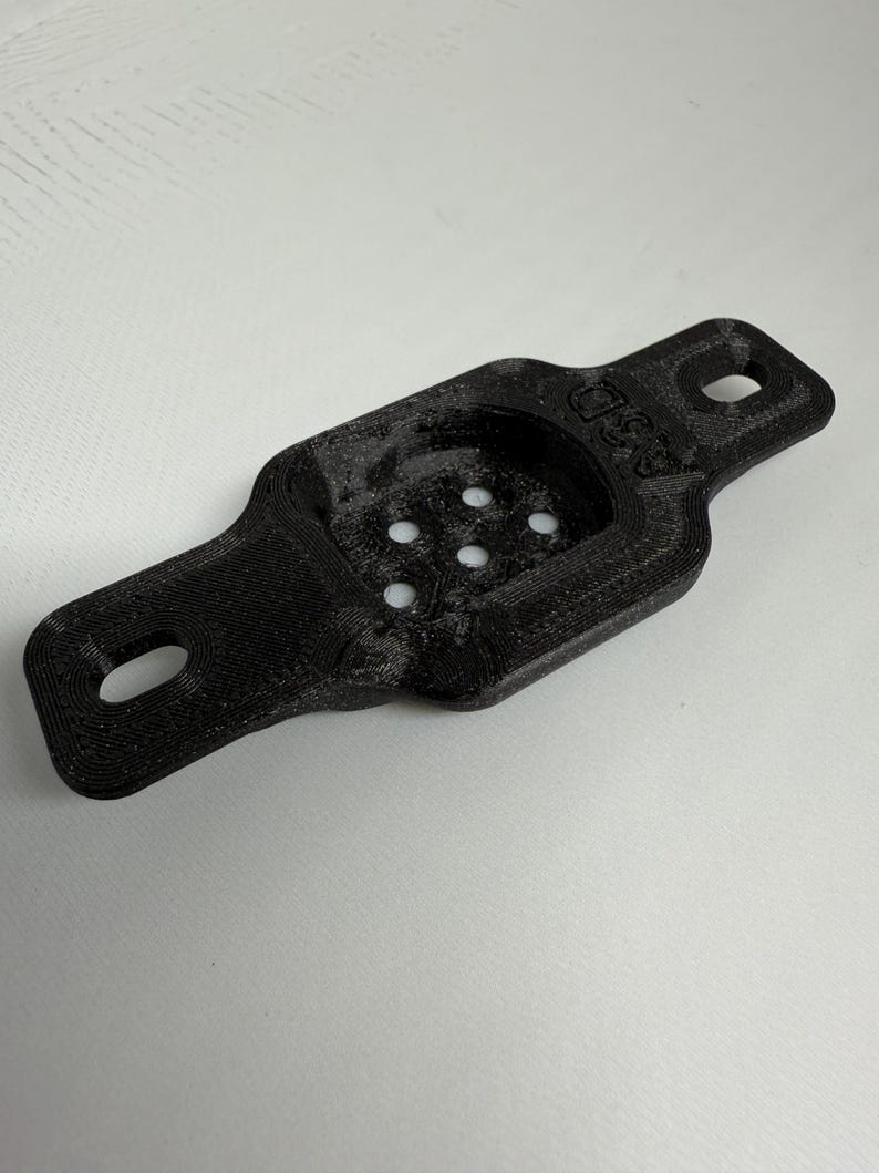 May include: A black 3D-printed bracket with a rectangular body and rounded ends. It features two holes at the ends and a recessed area with five smaller holes. The text "ASD" is visible on the side.