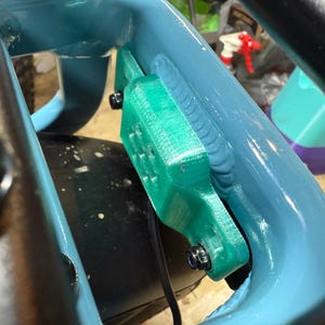 May include: Close-up of a teal-colored bicycle frame with a bright green component attached. The green piece is secured with black bolts. A black cable runs along the frame. The background is blurred.