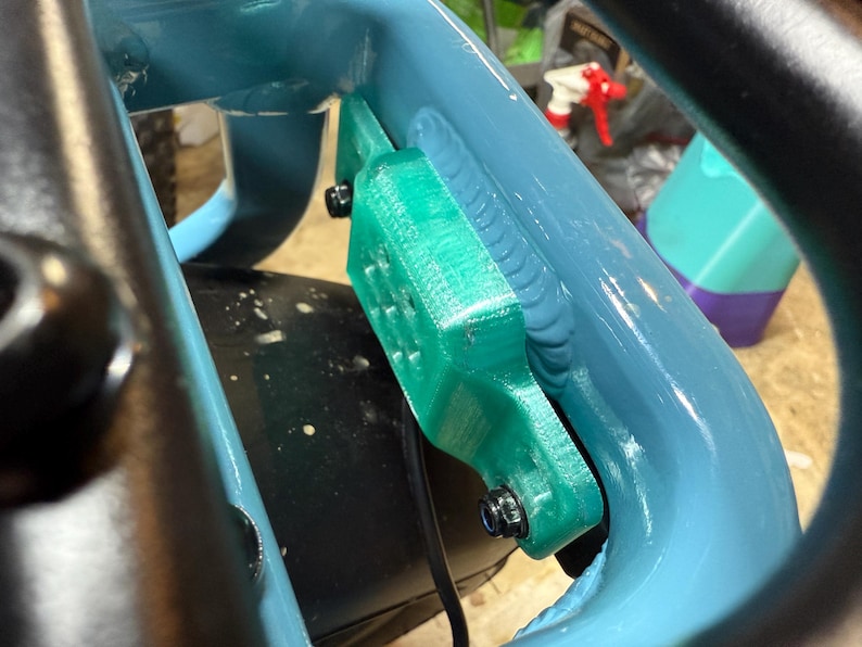 May include: Close-up of a teal-colored bicycle frame with a bright green component attached. The green piece is secured with black bolts. A black cable runs along the frame. The background is blurred.