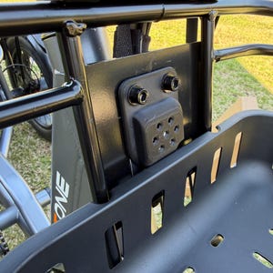 May include: Close-up of a black cargo basket attached to a bicycle. The basket is made of a durable, slotted material. The mounting hardware is visible, including a black bracket and bolts. The bicycle frame is gray with the word "ONE" printed on the side.