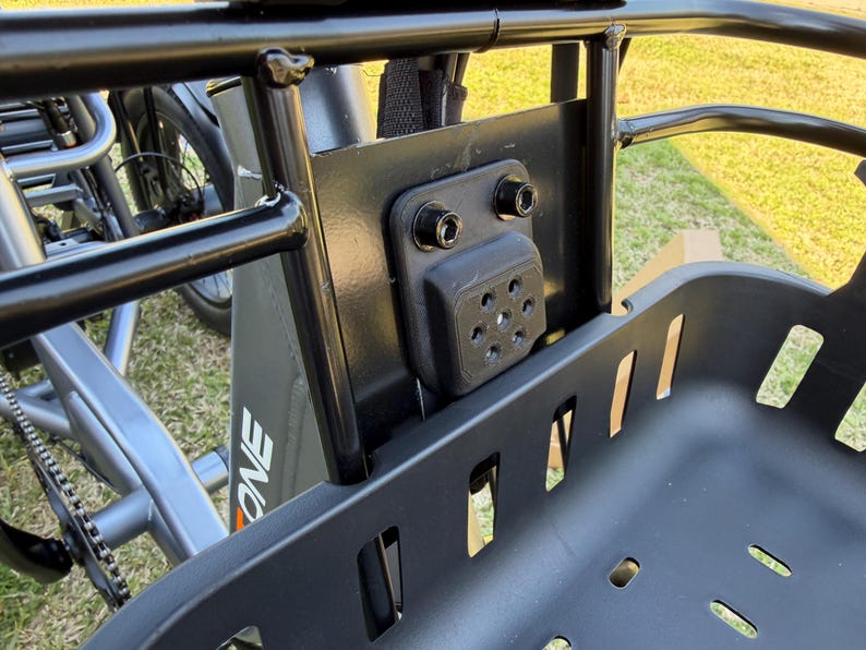 May include: Close-up of a black cargo basket attached to a bicycle. The basket is made of a durable, slotted material. The mounting hardware is visible, including a black bracket and bolts. The bicycle frame is gray with the word "ONE" printed on the side.