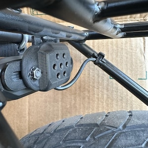 May include: Close-up of a black, 3D-printed component attached to a black metal frame, secured with a bolt. The component has multiple circular openings. A black tire is visible in the foreground, with a black wire secured with a zip tie.