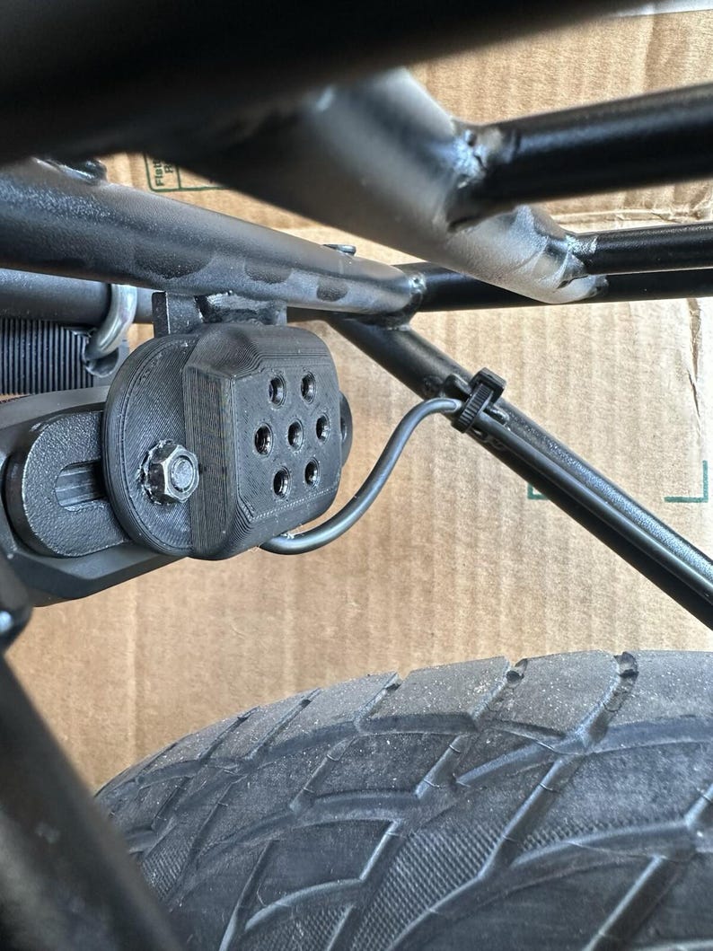 May include: Close-up of a black, 3D-printed component attached to a black metal frame, secured with a bolt. The component has multiple circular openings. A black tire is visible in the foreground, with a black wire secured with a zip tie.