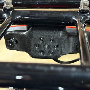 May include: Close-up of a black, 3D-printed component attached to a black metal frame. The component has multiple circular holes and is secured with bolts. A red light is visible behind the component.