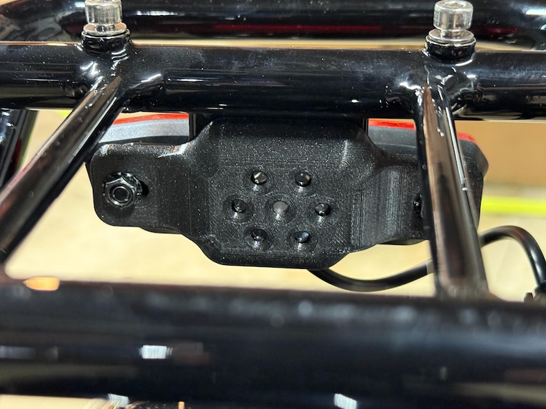 May include: Close-up of a black, 3D-printed component attached to a black metal frame. The component has multiple circular holes and is secured with bolts. A red light is visible behind the component.