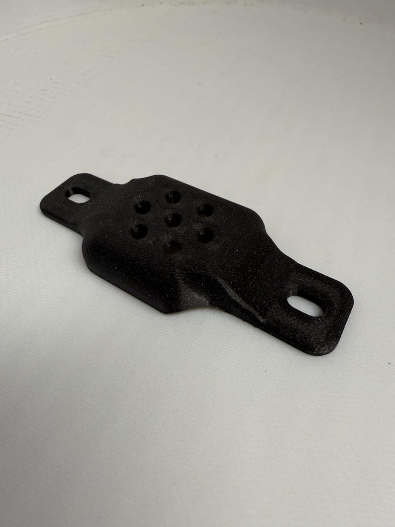 May include: A black, 3D-printed object with a rectangular shape and rounded edges. It features two holes on the ends and several smaller holes in the center. The object is likely a component or part of a larger assembly.