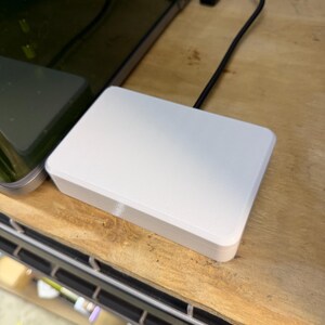 May include: A white rectangular electronic device with rounded edges, connected to a black cable. The device is resting on a wooden surface, with a dark monitor and other equipment visible in the background.