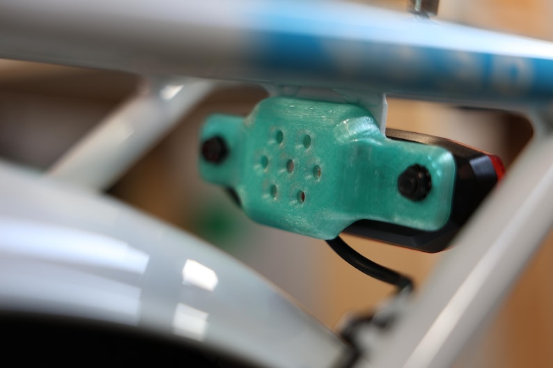 May include: Close-up of a bicycle tail light assembly. The light is black and red, mounted on a teal-colored bracket with multiple holes. The bracket is attached to a white bicycle frame with a blue accent.