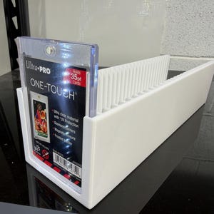 May include: A white card holder with dividers, holding a clear Ultra Pro One-Touch card case. The card case has the text "Ultra PRO ONE-TOUCH" and "35pt Cards". The holder is designed to organize and display trading cards.