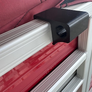 May include: A black, right-angled bracket with a circular opening, mounted on a red and silver metal surface. The bracket is positioned against a burgundy fabric background, suggesting a possible attachment point.