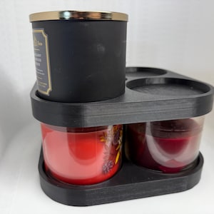 May include: Black candle holder with three circular openings for candles. The holder has a black candle with a gold lid and two clear glass candles with red and purple wax inside.