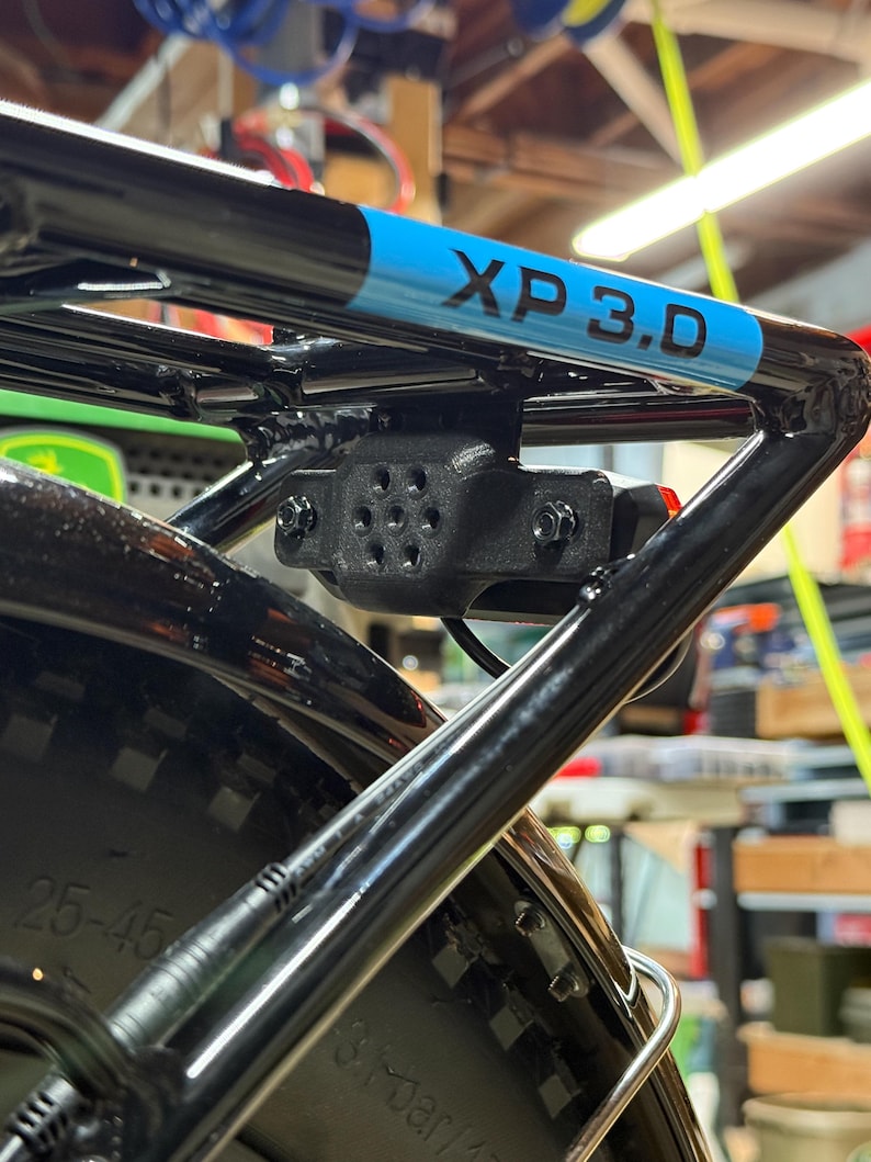 May include: Black bicycle rack with a blue and black stripe that reads "XP 3.D". The rack is mounted on a black tire. A black light is attached to the rack.