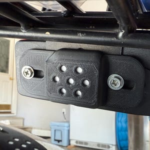 May include: Black plastic mounting bracket with seven circular holes and two silver screws, attached to a black metal frame. The bracket appears to be 3D-printed and is mounted on a vehicle.