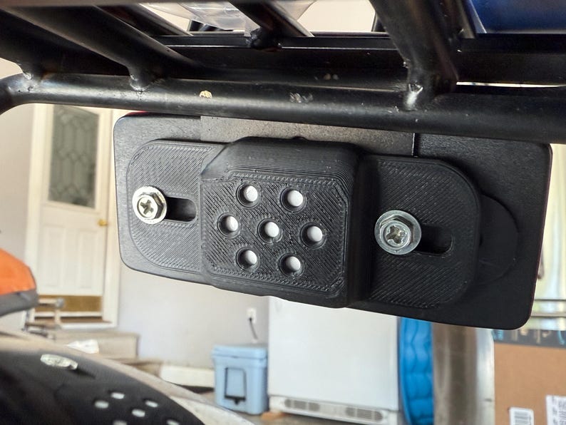 May include: Black plastic mounting bracket with seven circular holes and two silver screws, attached to a black metal frame. The bracket appears to be 3D-printed and is mounted on a vehicle.