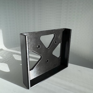 May include: A black, rectangular bracket with a textured surface. The bracket features a cut-out design with triangular and circular openings. The object is positioned at an angle, showcasing its depth and form. The background is a soft, neutral color.