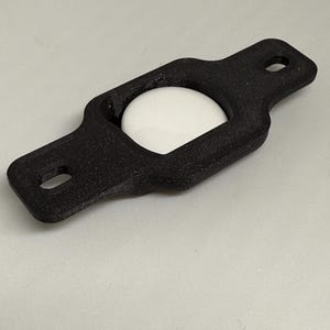 May include: A black, rectangular holder with rounded edges and two holes at the ends. A white, spherical object is nestled within the holder's center. The holder appears to be made of a textured material.