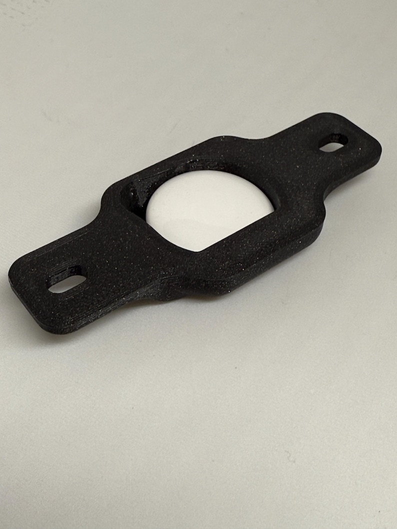 May include: A black, rectangular holder with rounded edges and two holes at the ends. A white, spherical object is nestled within the holder's center. The holder appears to be made of a textured material.