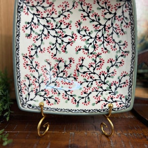 May include: A square ceramic serving dish with a floral pattern in shades of red, green, and black. The dish is on a gold metal stand.